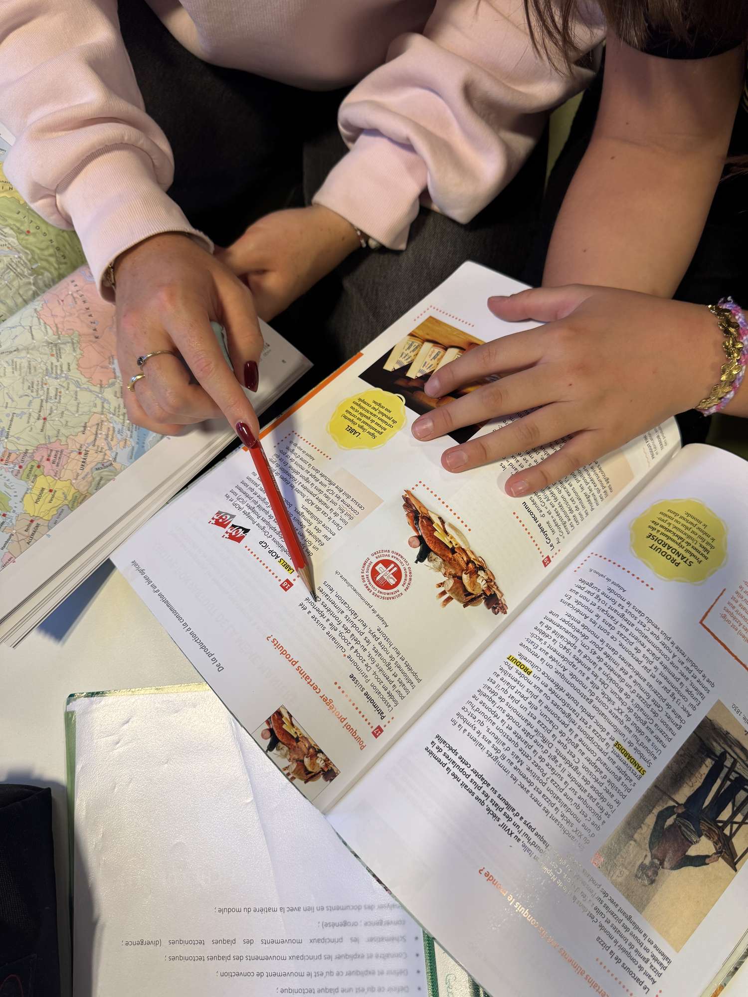 Vue rapprochée de deux paires de mains au-dessus d’un manuel de géographie ouvert : une tutrice désigne un passage avec un crayon rouge, l’élève pose la main sur la page, un atlas ouvert à côté.