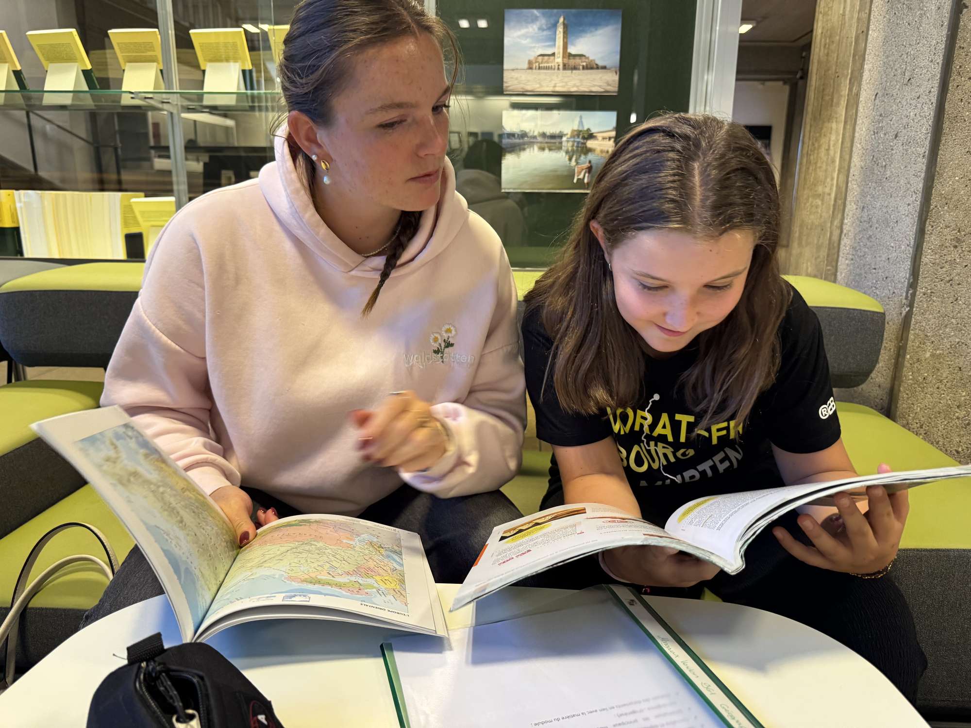 Assise sur un canapé vert dans une bibliothèque, une tutrice observe avec attention une élève qui lit son manuel, un atlas ouvert devant elles.