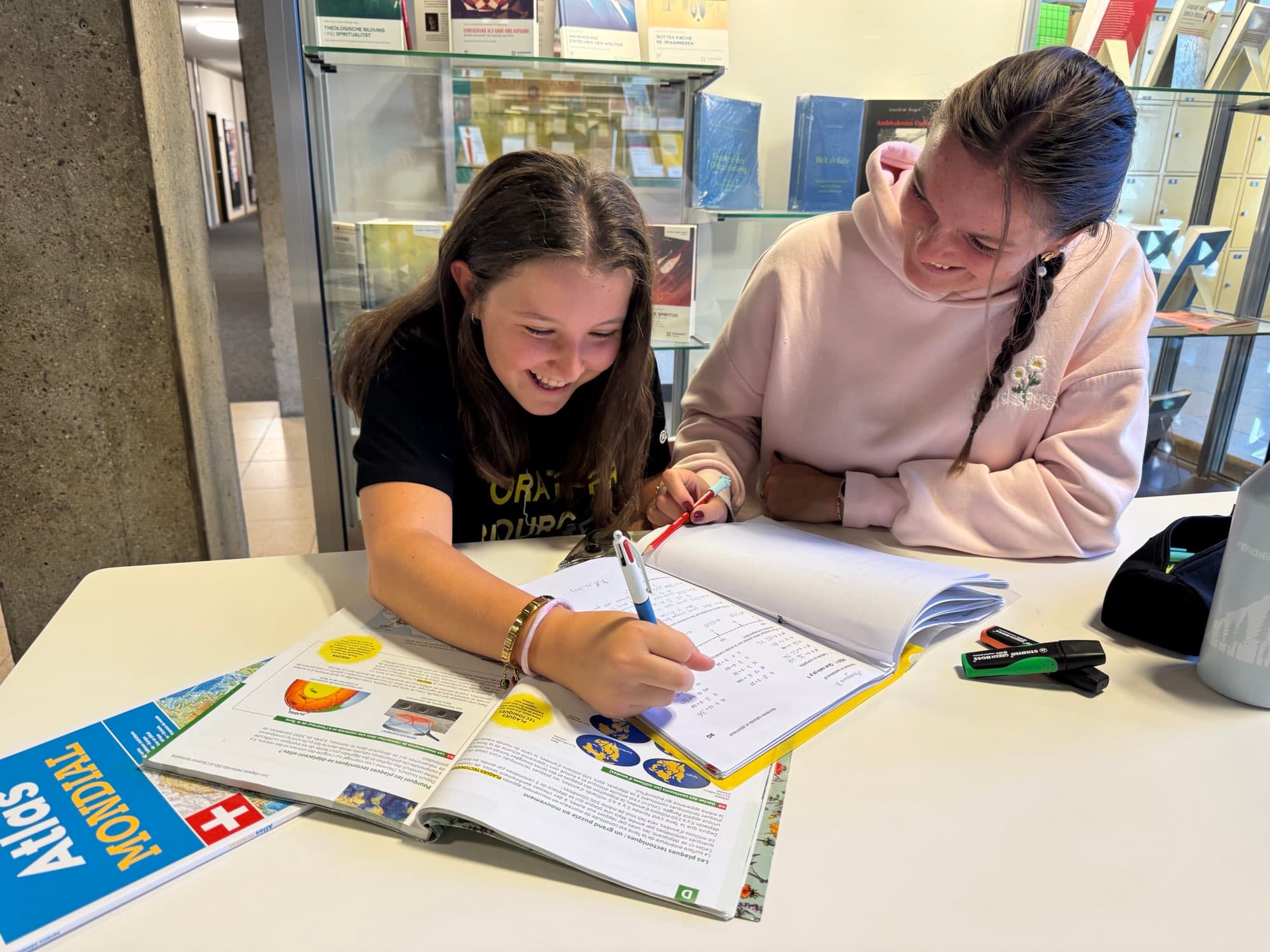 Une élève rit en remplissant son cahier à une table de bibliothèque, à côté de sa tutrice qui sourit en suivant son travail, un atlas ouvert devant elles.