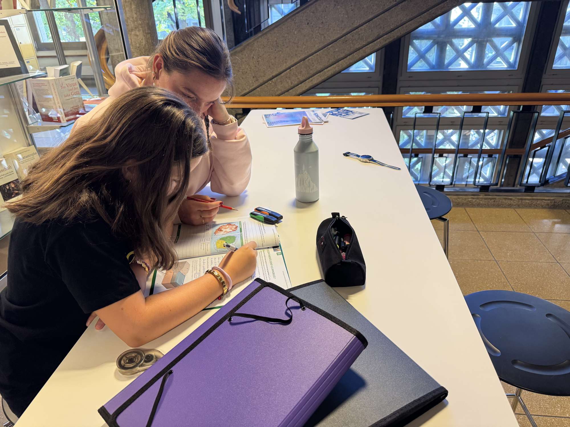 Une élève écrit dans son livre à une grande table blanche de bibliothèque, tandis que sa tutrice, accoudée à côté d’elle, suit son travail un crayon à la main.